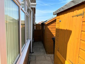 A narrow outdoor space with a wooden fence, a window with blinds, and a concrete pathway. - 2-bed Bungalow, Sleeps 6, Parking & Pet Friendly (Skegness)