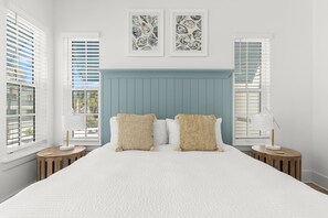 A bedroom with a large bed, two bedside tables, a headboard, and two windows with blinds.