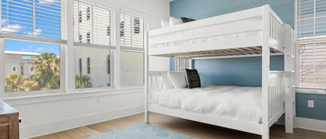 Habitación con literas, barandillas blancas, una pared de acento azul y ventanas grandes con persianas.