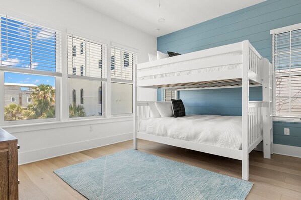 A bunk bed room with white railings, a blue accent wall, and large windows with blinds.