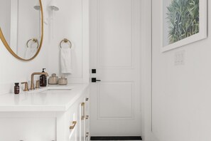 A white bathroom with a gold-framed mirror, a white sink with gold fixtures, and a framed plant on the wall.
