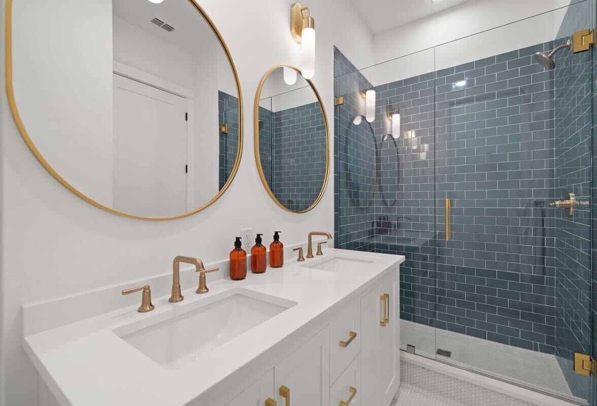 Une salle de bain avec un meuble-lavabo blanc, deux grands miroirs, une douche aux carreaux bleus et des accessoires dorés.