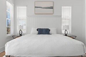 Ein Schlafzimmer mit einem großen Bett, weißen Wänden und zwei Fenstern mit Jalousien.