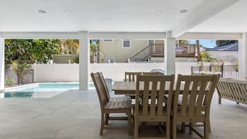 A covered patio area with a pool, wooden furniture, and a view of a residential building.