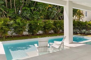 A swimming pool with two white lounge chairs, surrounded by lush greenery and a white building in the background. - Onyx House at The Gemma (Clearwater)