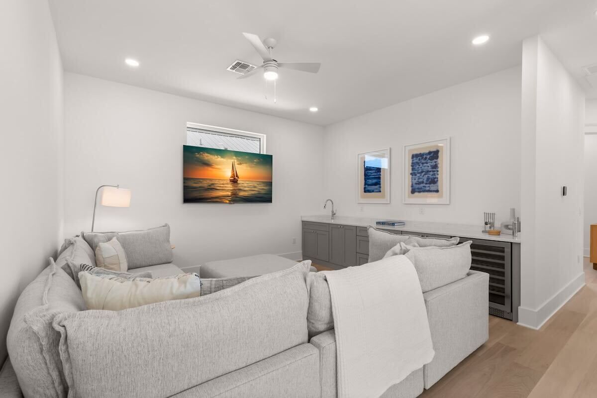 A modern living room with a sofa, a ceiling fan, a painting of a sunset, and a kitchen area with cabinets and a sink.