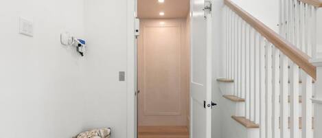 Un pasillo con piso de madera, pared blanca, puerta blanca y escalera de madera con peldaños.