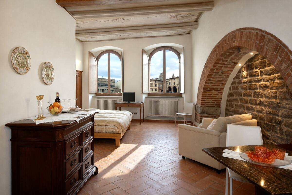 Una habitación con cama, cómoda, mesa con fruta, dos ventanas con vistas y un arco de piedra.