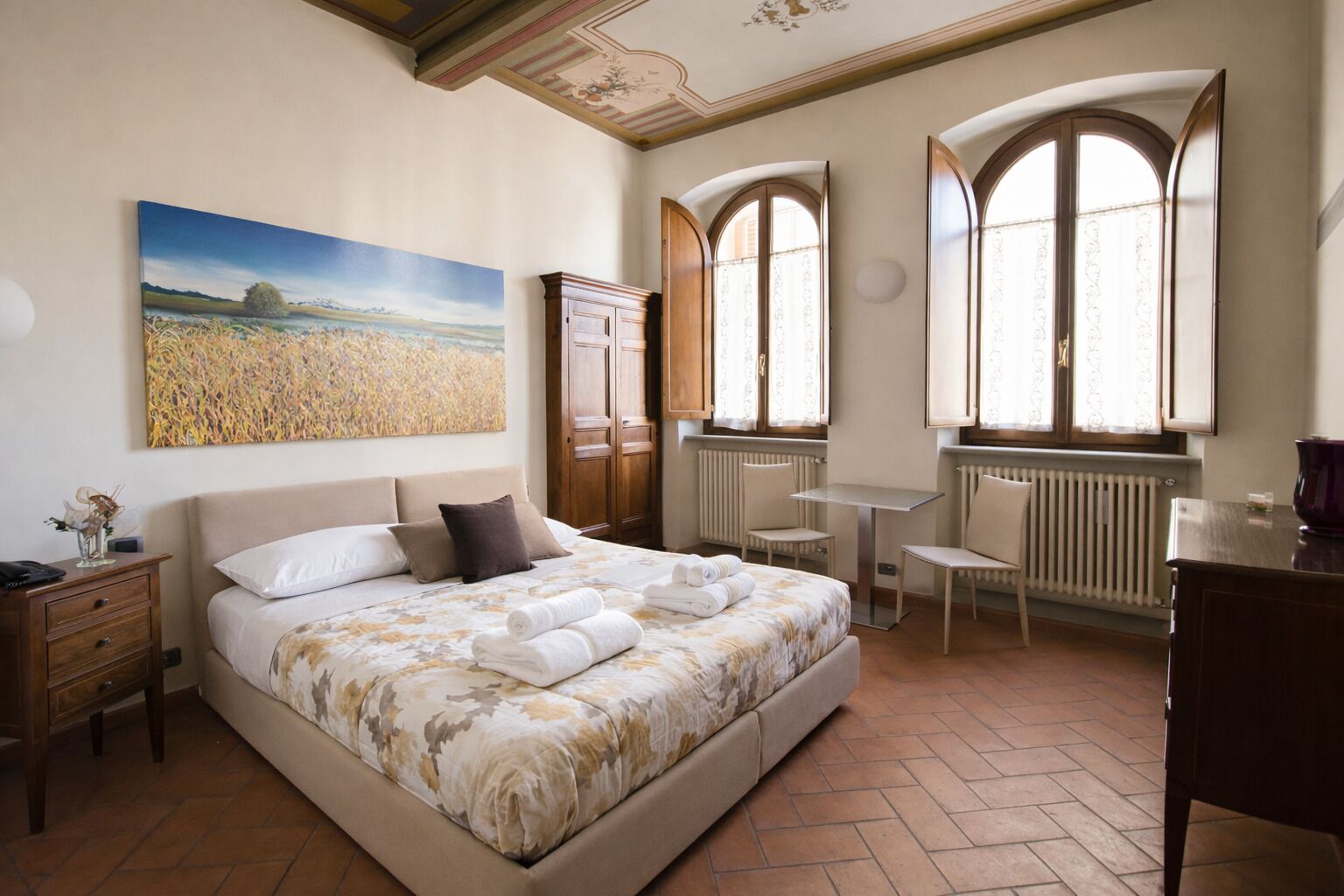 A bedroom with a large bed, a desk, two chairs, a radiator, and a window with curtains.