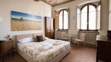 A bedroom with a large bed, a desk, two chairs, a radiator, and a window with curtains.