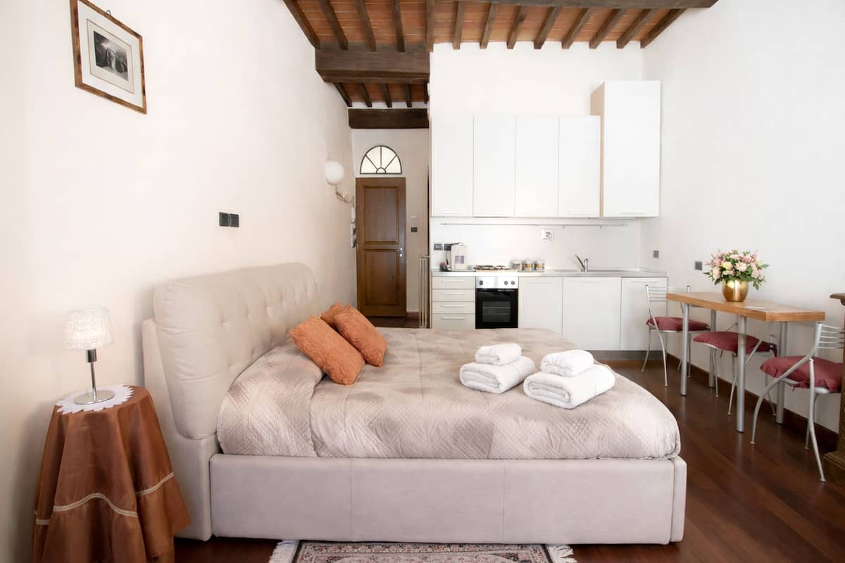 Un dormitorio con cama, mesita de noche, lámpara, una pequeña mesa con un florero y una kitchenette al fondo.
