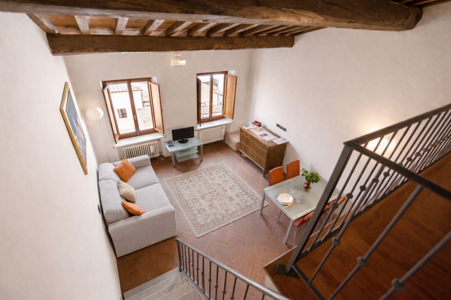 A modern living room with a sofa, a dining table, and a TV. There are windows and a staircase.