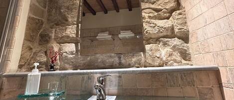 Un baño con pared de piedra, lavabo de vidrio y vigas de madera en el techo.