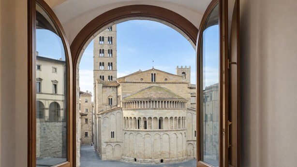 Vista a través de un ventanal de arco de un edificio histórico, con una torre de campanas, un patio interior y arquitectura circundante.