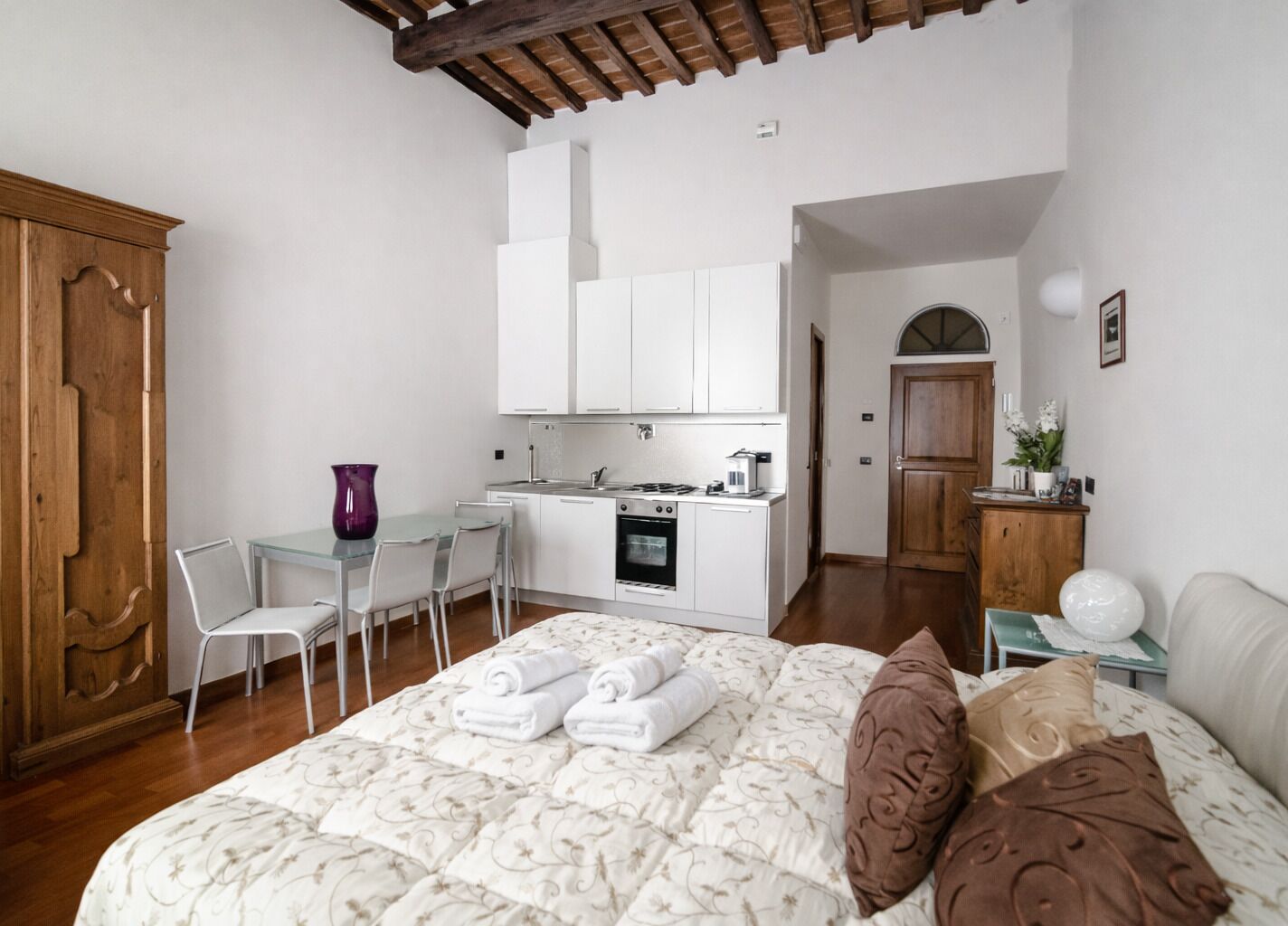 A compact hotel room with a bed, a small kitchenette, a dining area, and a wooden wardrobe.