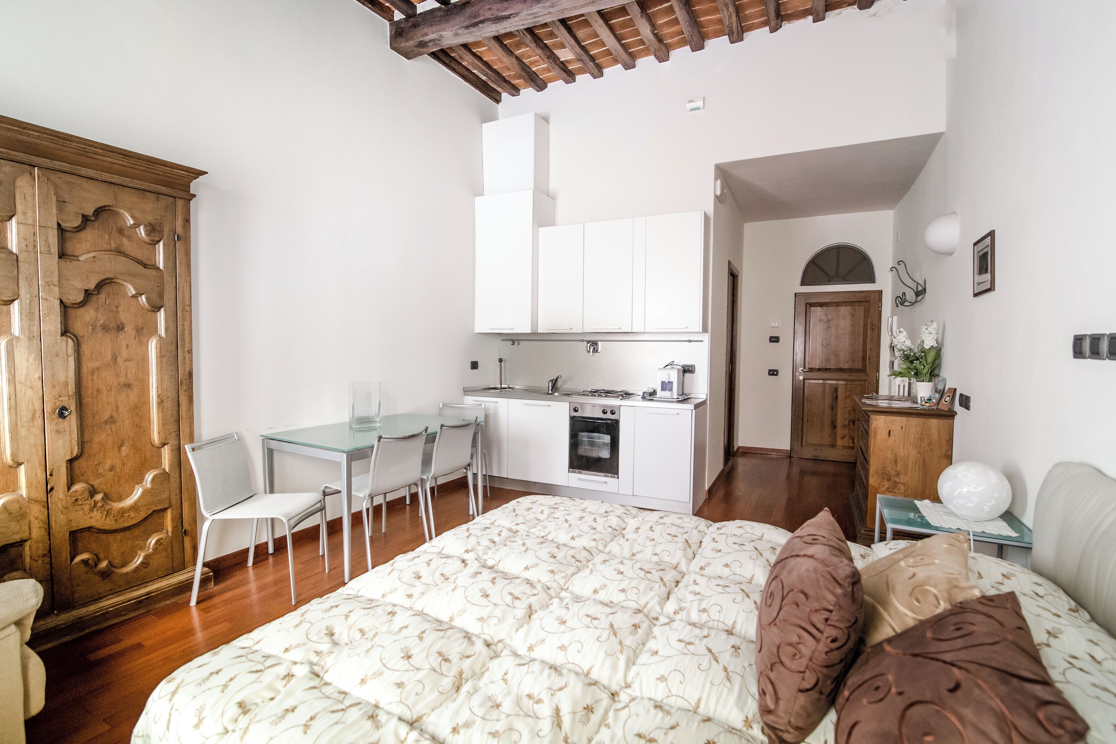 Quarto com uma cama grande, um guarda-roupa de madeira, uma pequena cozinha com armários brancos e uma área de jantar com cadeiras e mesa.