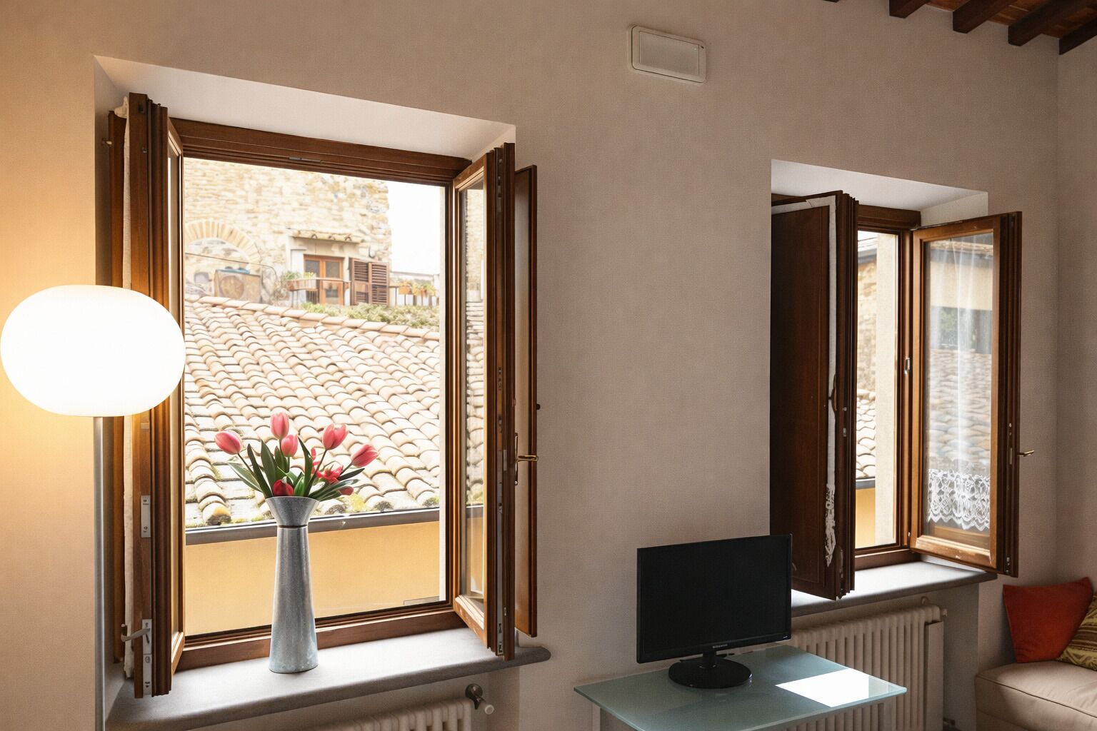 A room with a window, a vase with flowers, a computer monitor, and a radiator.
