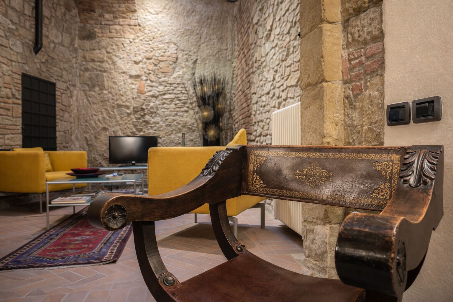 A stone-walled room with a wooden bench, a yellow sofa, and a television.