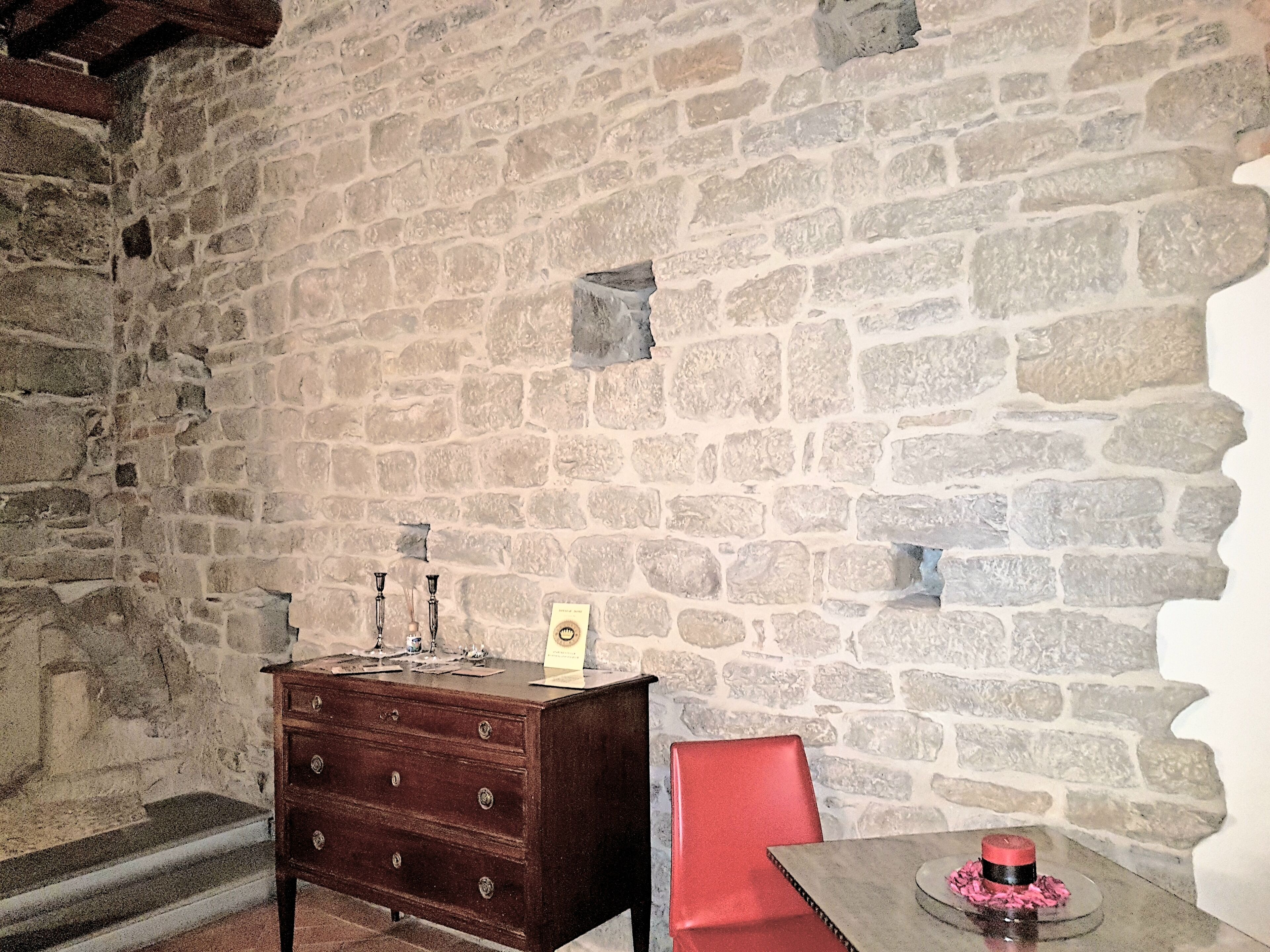 Una habitación con paredes de piedra, un aparador de madera, una silla roja y una mesa con un adorno.