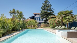 A rectangular swimming pool with a wooden deck in the foreground, surrounded by lush greenery and a fenced area. - KALIKO (Bidart)