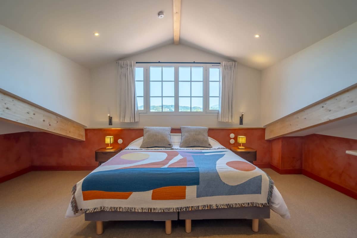 A bedroom with a large bed, two bedside tables with lamps, a window with curtains, and a sloped ceiling.