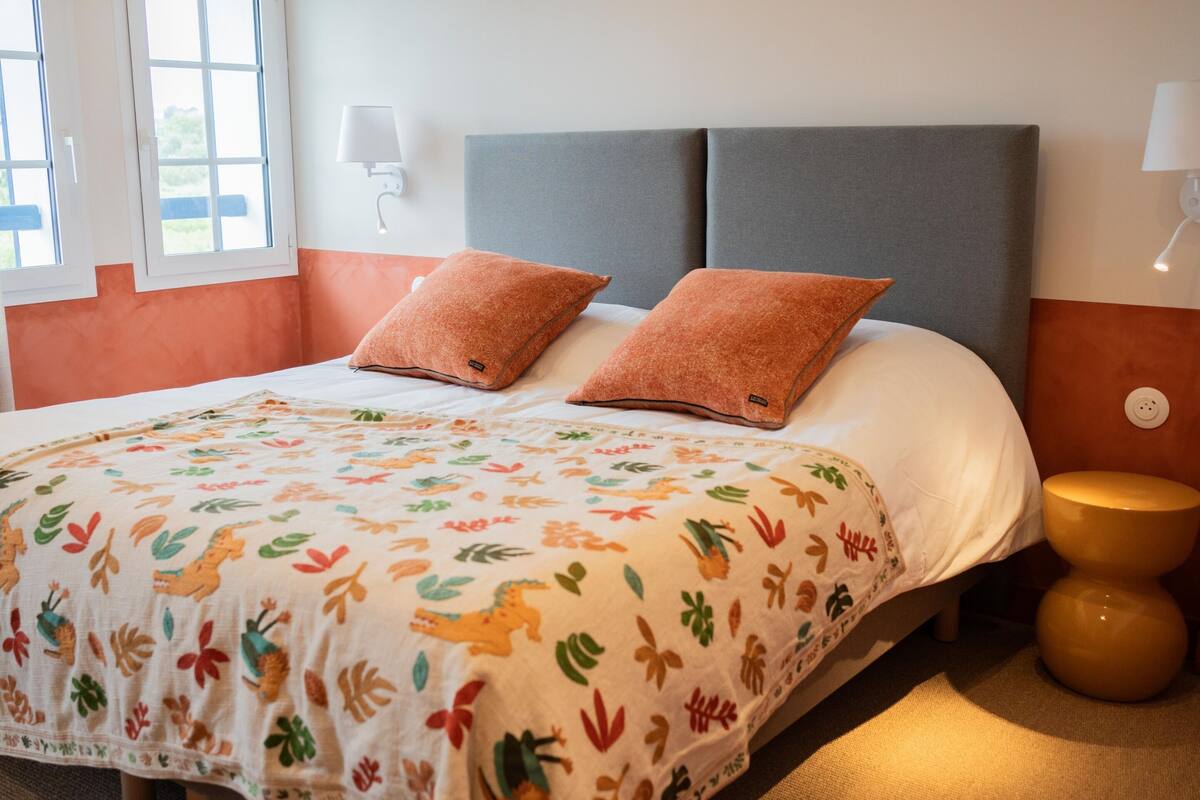 A neatly made bed with a colorful floral bedspread, a grey upholstered headboard, and a small round nightstand with a lamp.