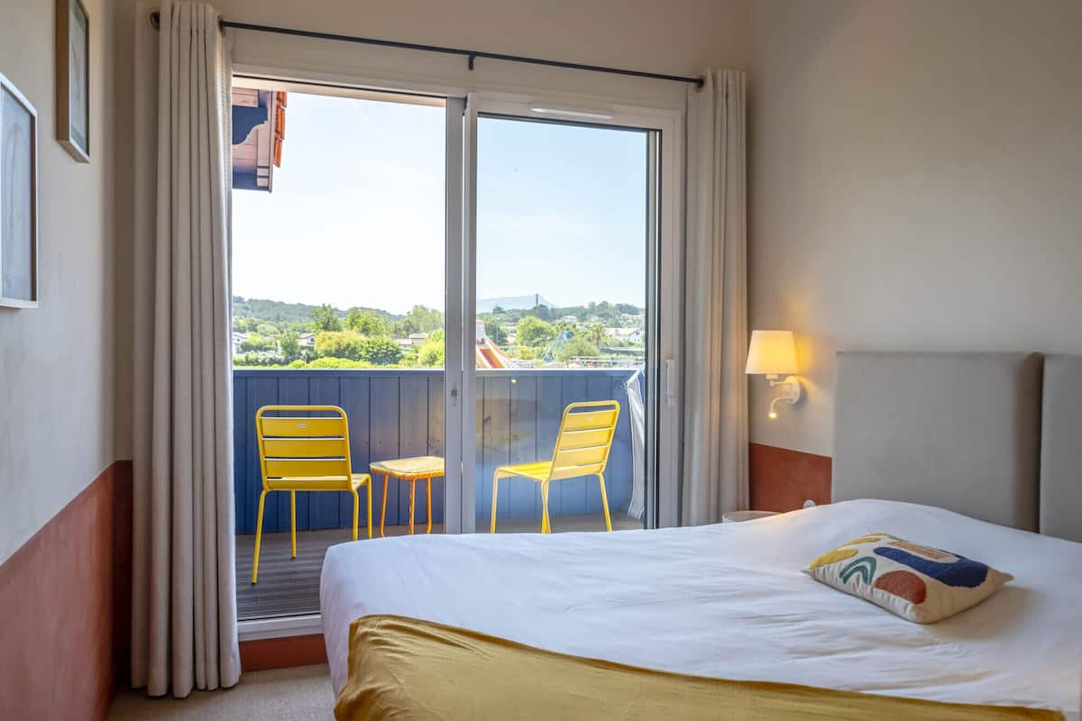A hotel room with a bed, two yellow chairs, a balcony with a view of greenery and buildings, and a wall-mounted lamp.