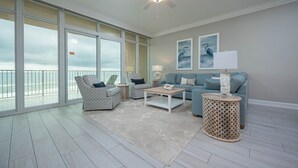 A spacious living room with large windows, a ceiling fan, a light-colored sofa, two armchairs, a coffee table, and a decorative lamp.