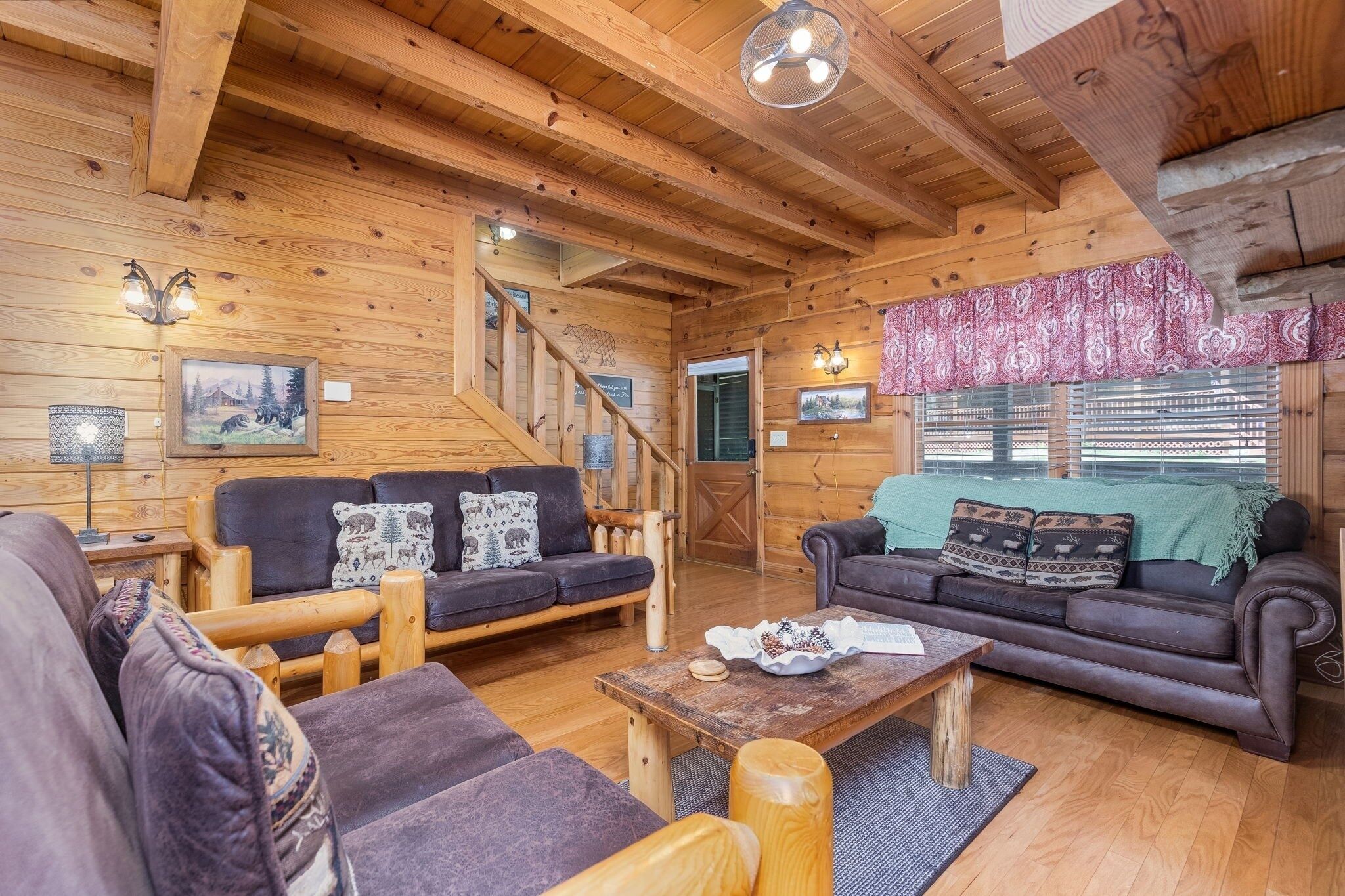 A cozy log cabin interior with wooden walls and ceilings, two sofas with patterned cushions, a wooden coffee table, and a staircase leading to a door.
