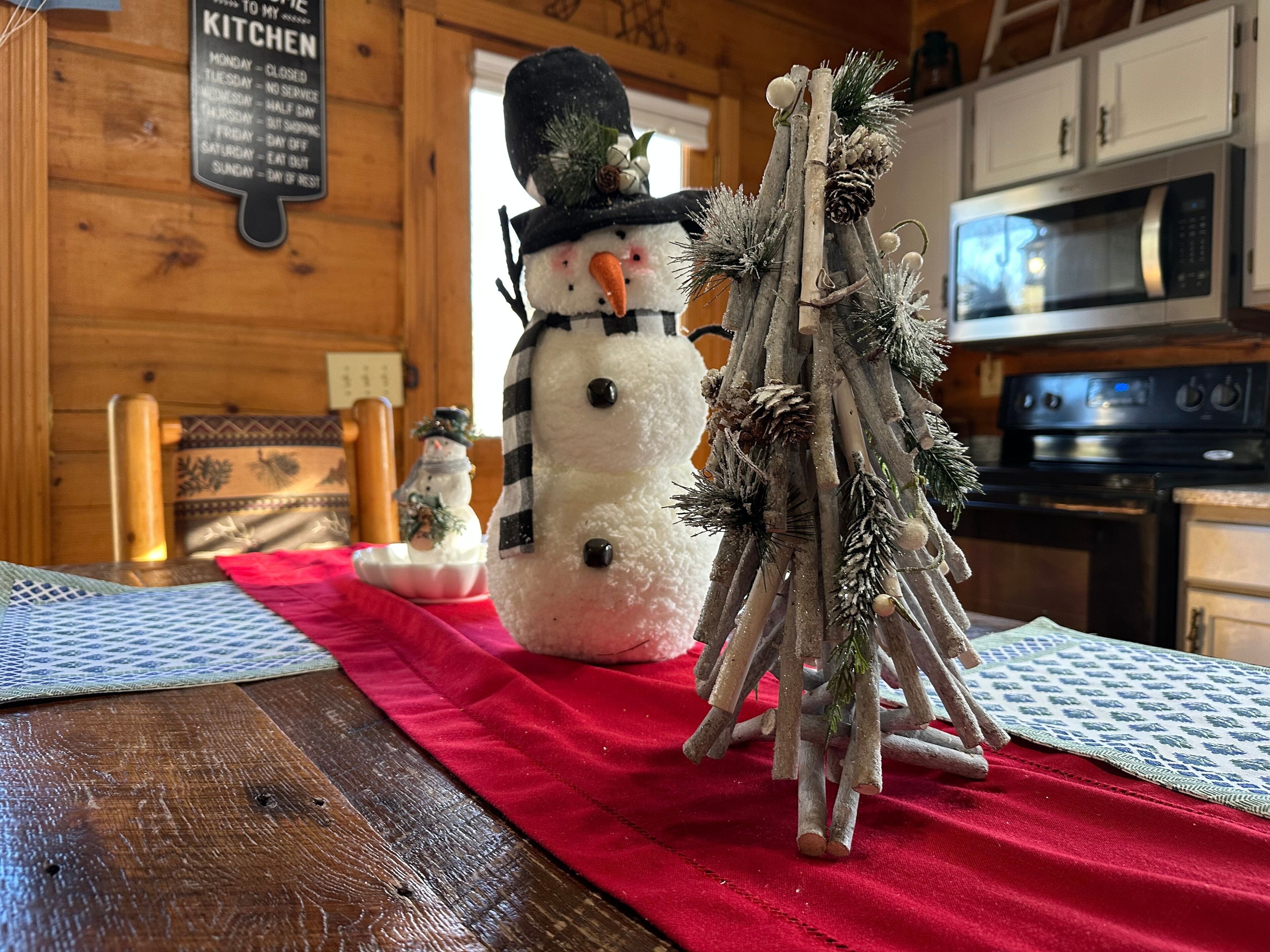 Interior de madera con zona de cocina, que incluye un microondas y estufa. Un mantel rojo cubre la mesa, y hay dos adornos de muñecos de nieve sobre ella.