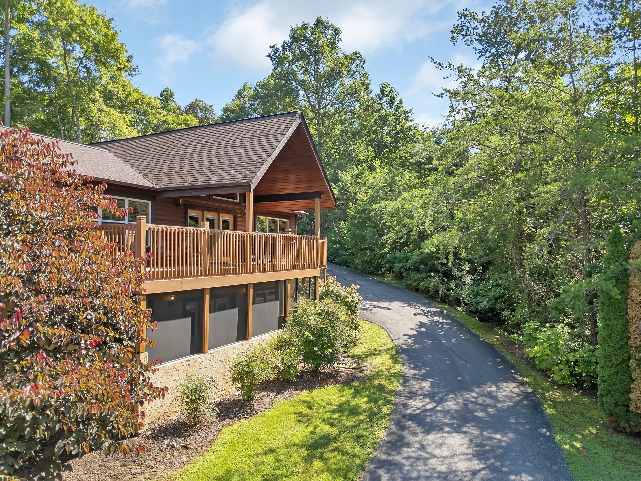 A wooden cabin with a balcony, surrounded by lush greenery and a winding driveway.