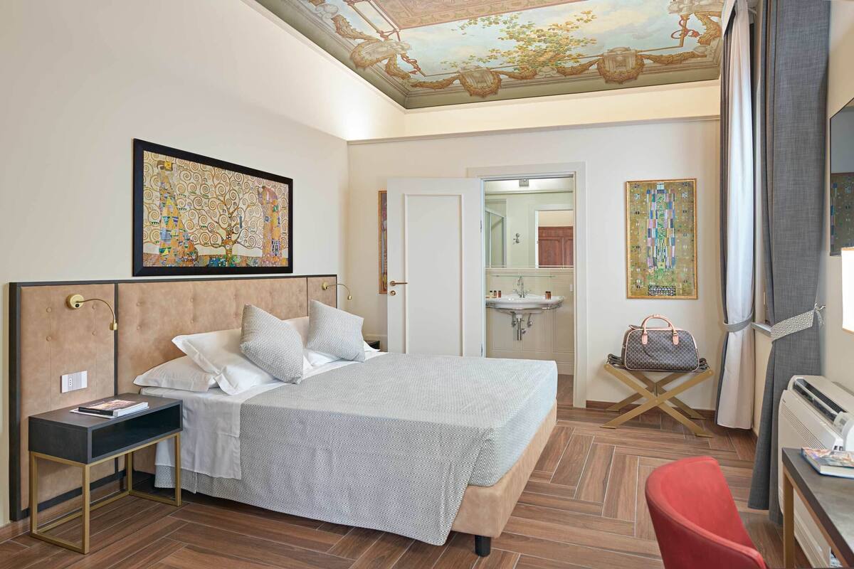A hotel room with a large bed, a bedside table, a red chair, a small table with a handbag, a bathroom with a sink and mirror, and a ceiling mural.