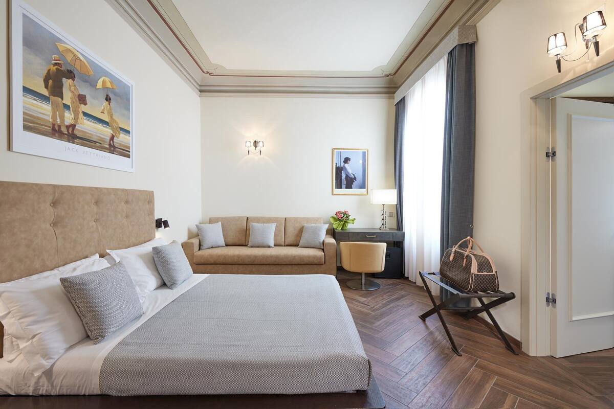 A hotel room with a large bed, a sofa, a desk with a lamp, and a chair. There is a painting on the wall and a door to the right.