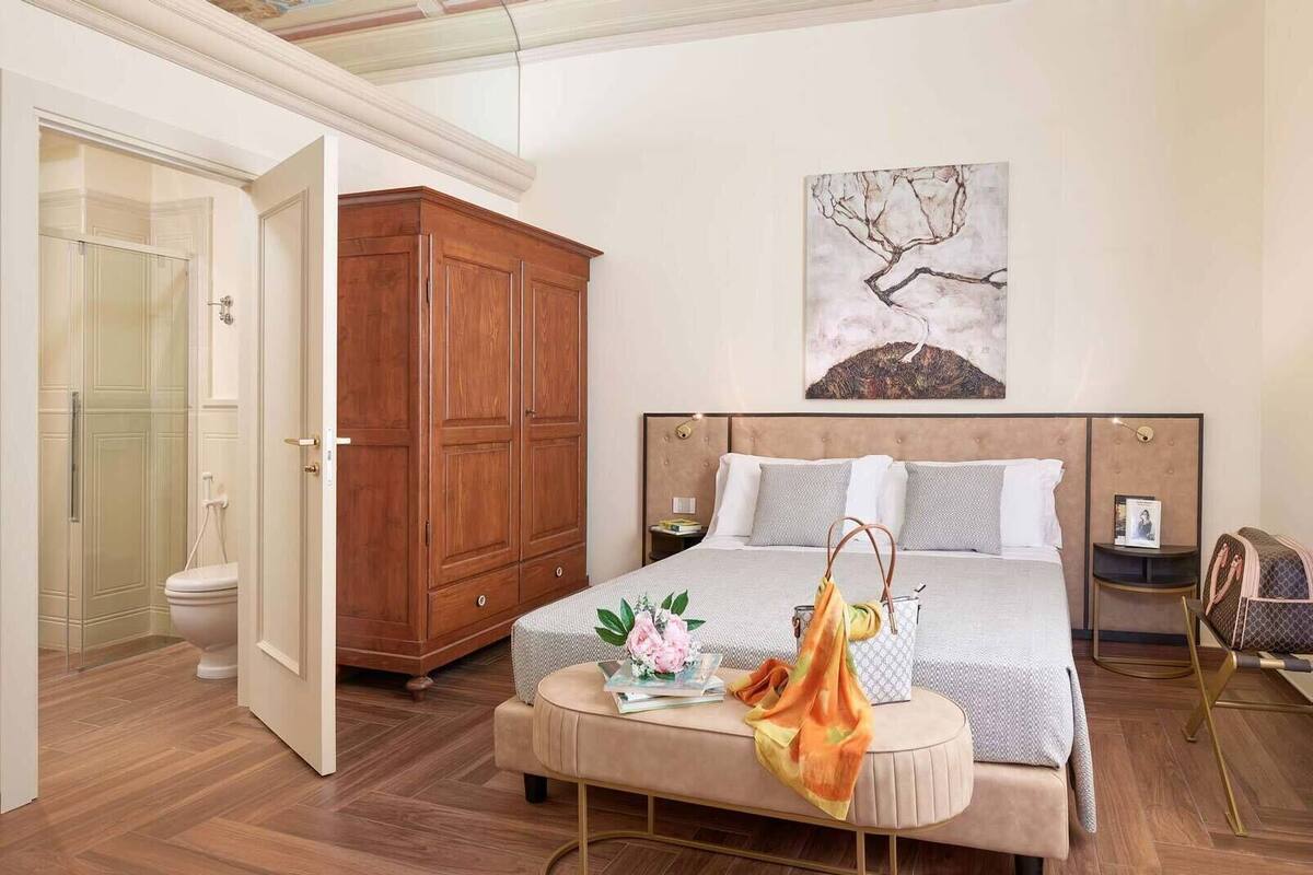 A bedroom with a large bed, a wooden wardrobe, a bathroom with a glass shower, and a painting on the wall.