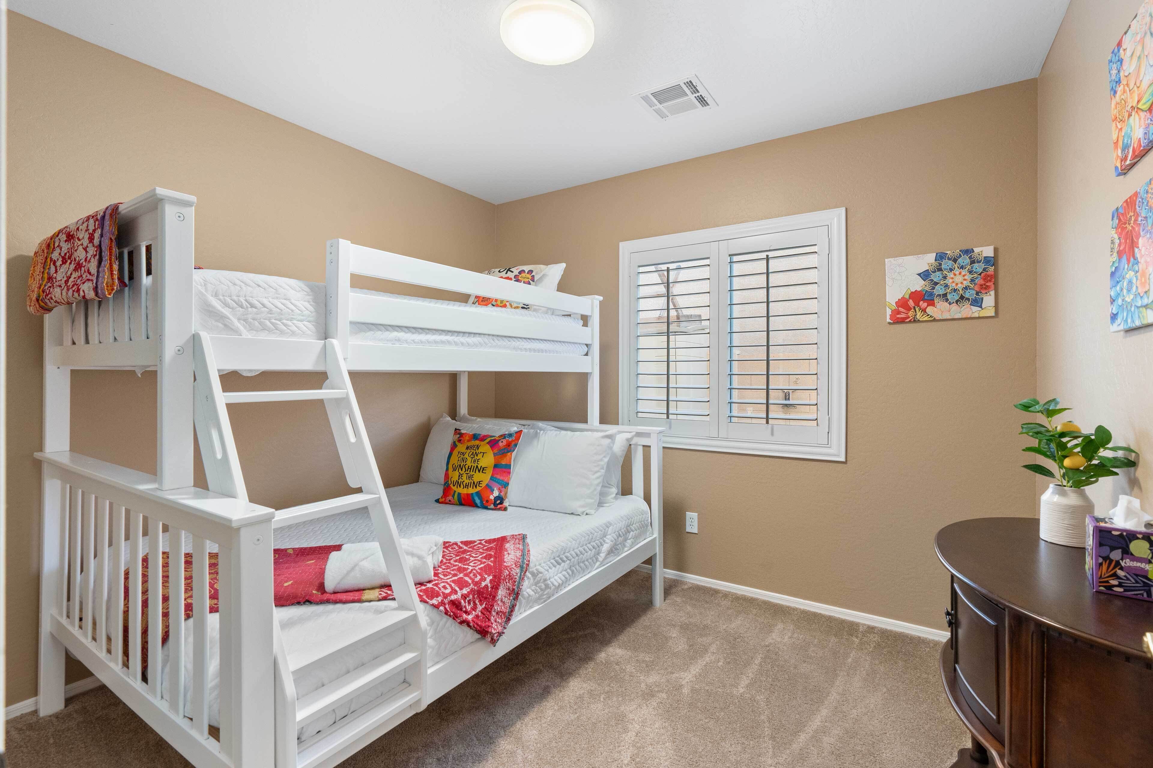 A bunk bed with a ladder, a window with blinds, and a small table with a vase and a box.