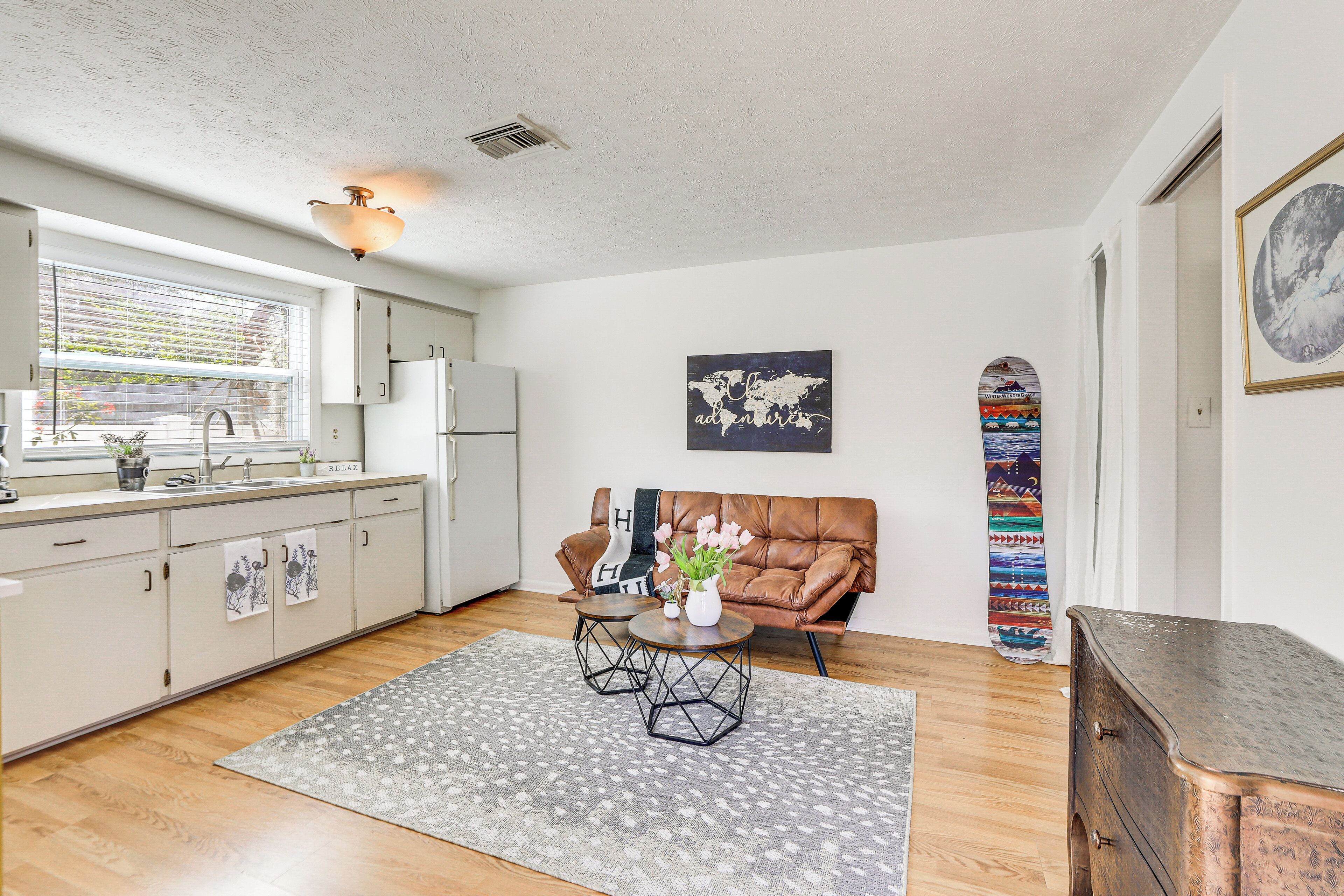 A modern kitchen with a window, white cabinets, a brown leather couch, and a skateboard leaning against the wall.