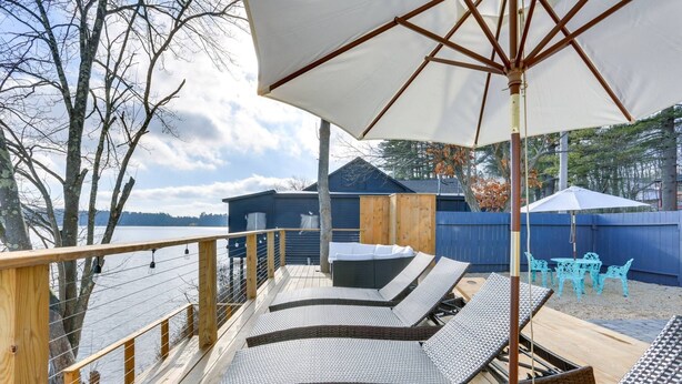 A deck with lounge chairs and an umbrella, overlooking a body of water and a building with a blue exterior. - Victorian Lake Oasis | Hot Tub | Private Lakefront (Damascus)