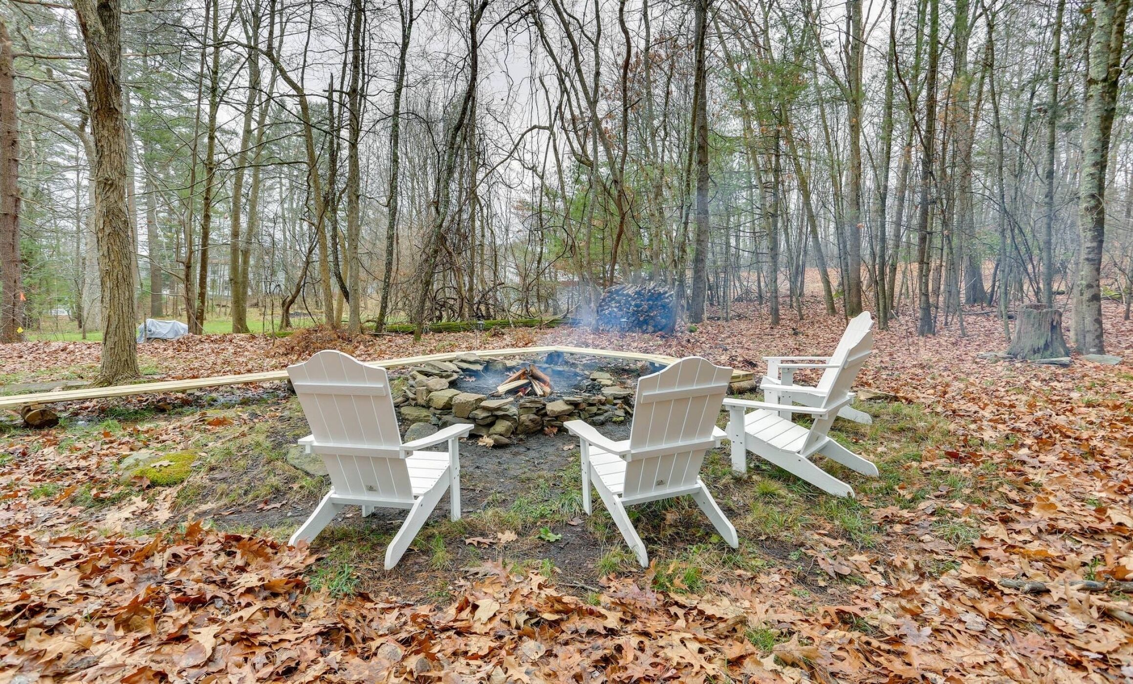 A forest setting with a fire pit and three Adirondack chairs.