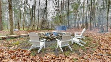 A forest setting with a fire pit and three Adirondack chairs.
