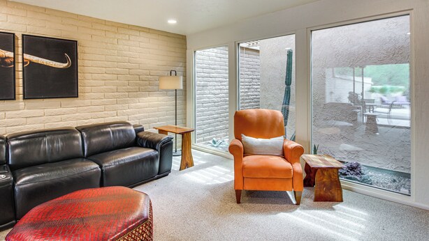 A modern living room with a black leather sofa, an orange armchair, and a red patterned ottoman. There is a wooden coffee table and a lamp on a side table. The room features a brick wall and large windows with a view of the outdoors. - Tucson Townhome w/ Mountain View, Pool Access! (Tucson)