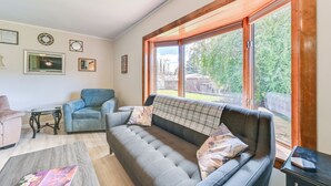 Un salon avec un canapé gris, un fauteuil bleu et une table basse. De grandes fenêtres offrent une vue sur les arbres et une clôture.