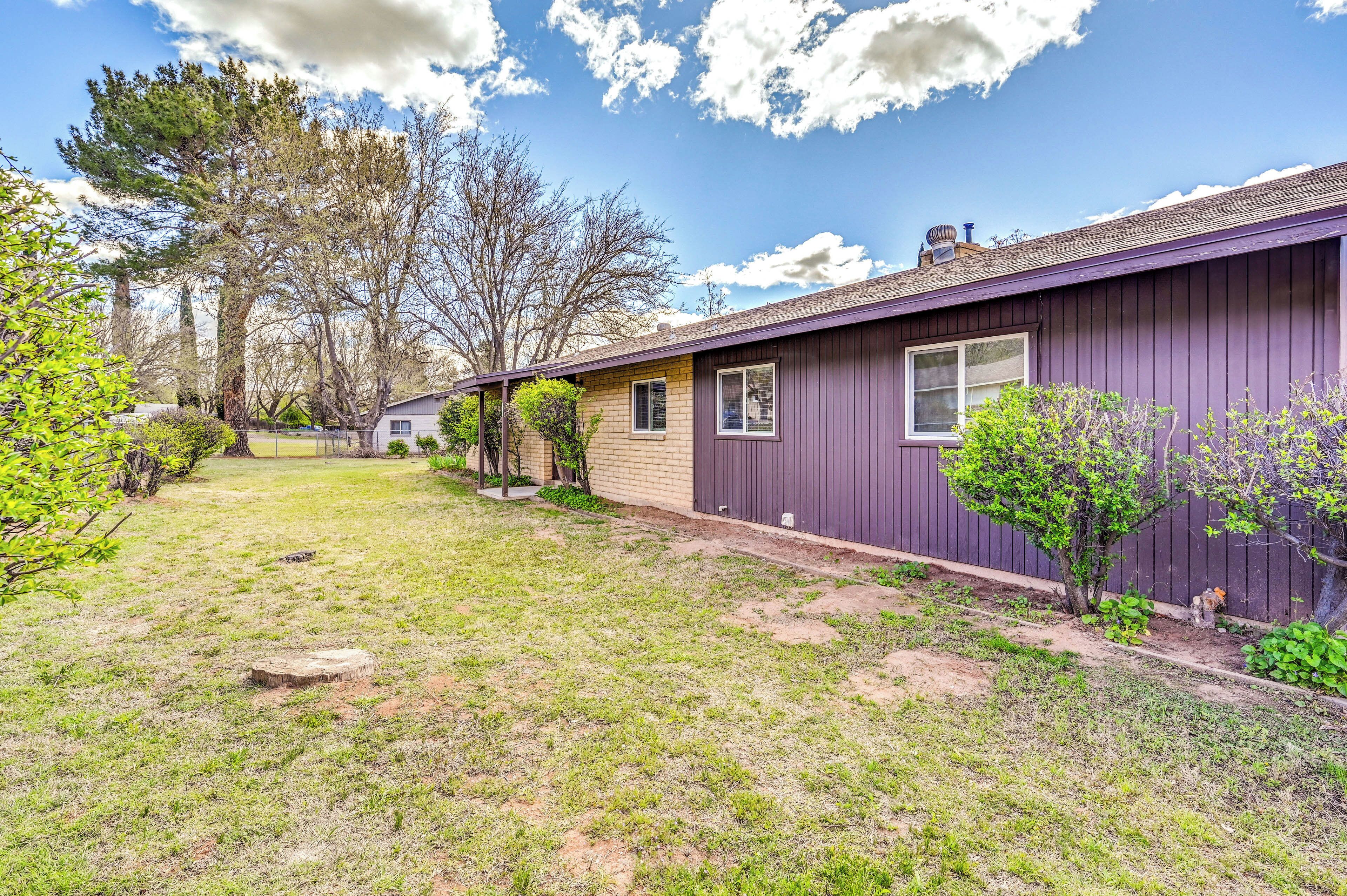 A single-story house with a purple exterior, a front yard with grass and small trees, and a clear blue sky with clouds.