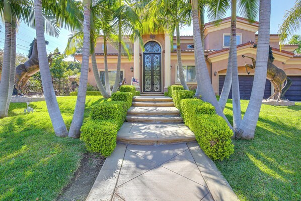 Stunning entrance with a central pathway leading to a gated door, surrounded by manicured hedges and palm trees.