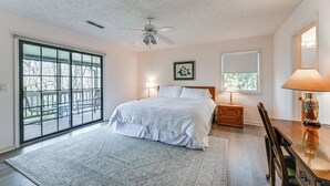 Un dormitorio con una cama grande, un ventilador de techo, una mesita de noche con lámpara, una silla de madera y vistas a través de un gran ventanal corredera.