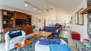 Amplia sala de estar con un sofá grande, una mesa de centro y un televisor. Se observa una cocina al fondo, con una mesa y sillas de comedor.
