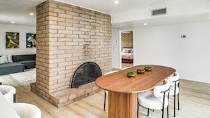 Uma sala de estar moderna com lareira de tijolos, uma mesa de jantar de madeira com cadeiras brancas e um sofá cinza.
