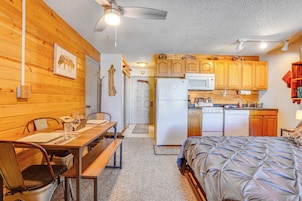 A compact room with a bed, a dining table with chairs, a kitchen area with wooden cabinets and appliances, and a ceiling fan.