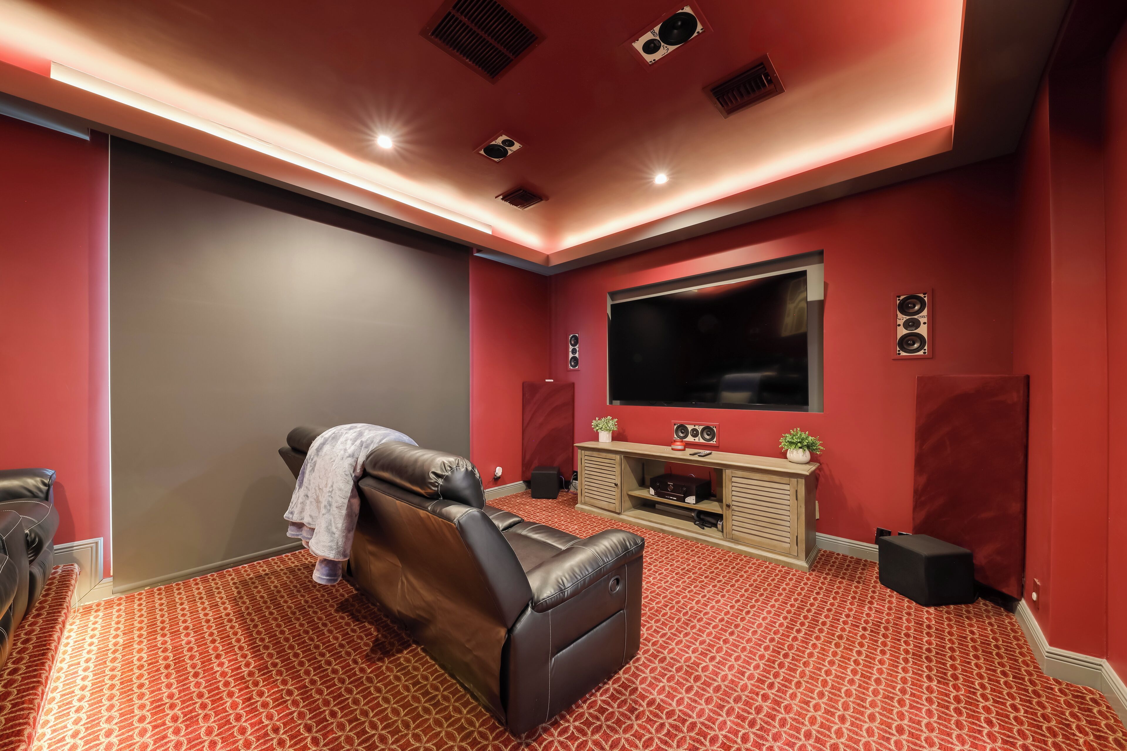Een bioscoopzaal in huis met een rood tapijt, leren fauteuil, televisie, speakers en een console tafel.