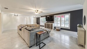 A spacious living room with a grey sectional sofa, a wooden coffee table, a ceiling fan, a flat-screen TV, and a large window with blinds. - Pool & Home Theater: Lighthouse Point Home! (Pompano Beach)