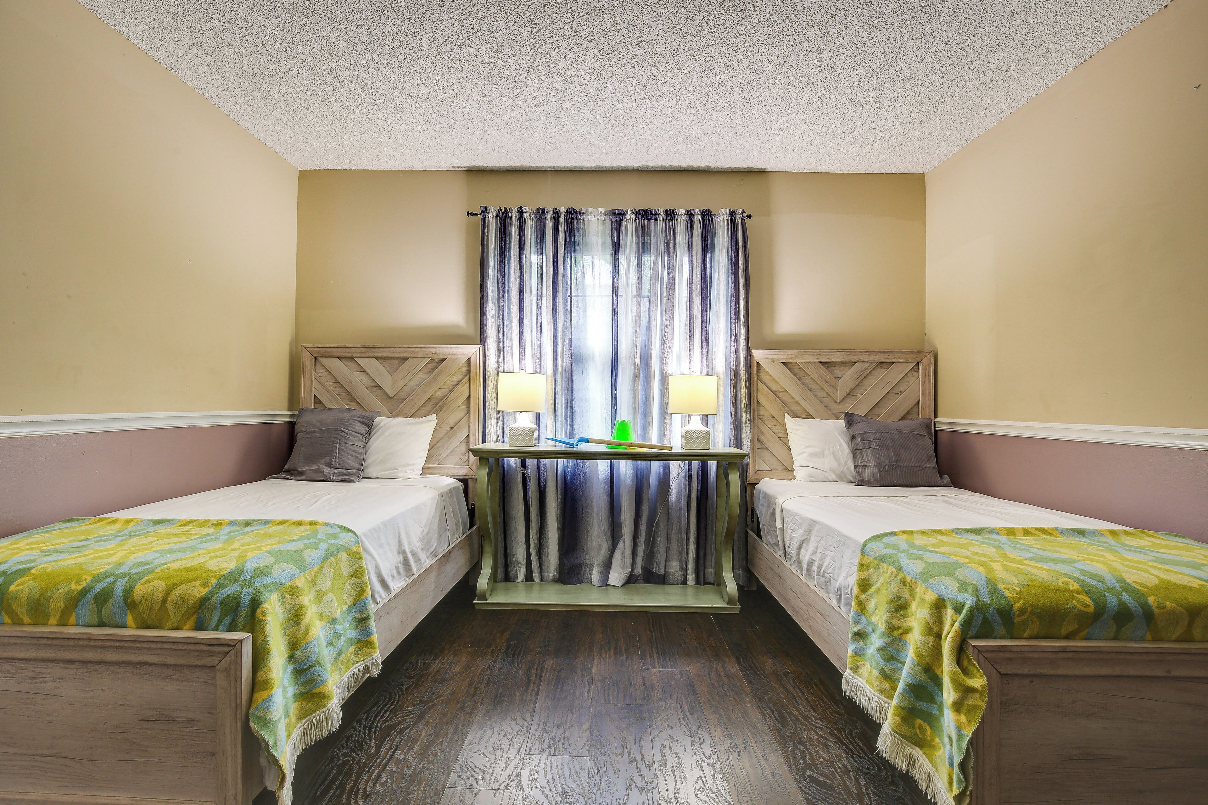 Quarto de hotel com duas camas de solteiro, uma mesinha de cabeceira de madeira com luminárias e vista através de cortinas leves.